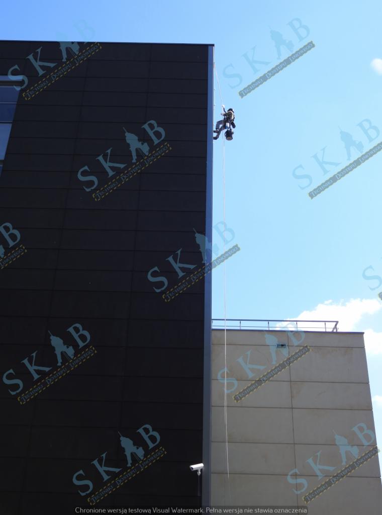 Pracownik na linie, zabezpieczony uprzężą, wykonuje prace na wysokości na elewacji nowoczesnego budynku z ciemnymi panelami, na tle błękitnego nieba.