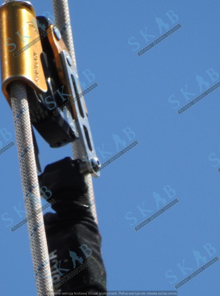 Przyrząd asekuracyjny na linie, używany podczas prac na wysokościach, w tle błękitne niebo.