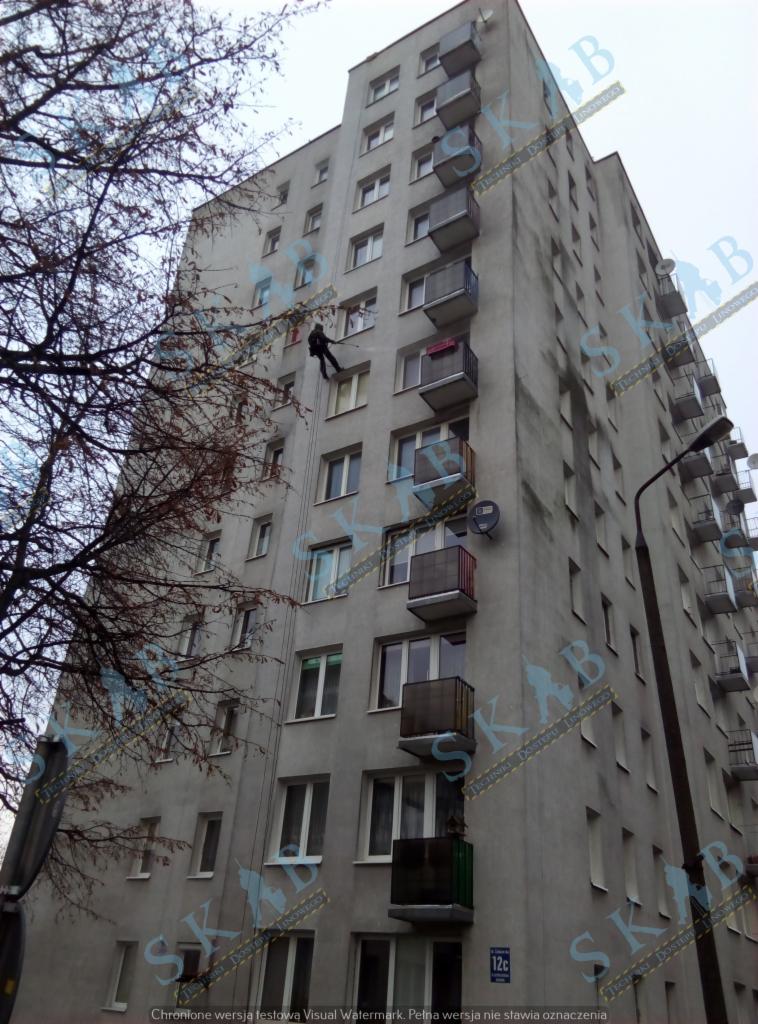 Pracownik na linach podczas prac na elewacji szarego bloku mieszkalnego, widoczne balkony i drzewo bez liści.