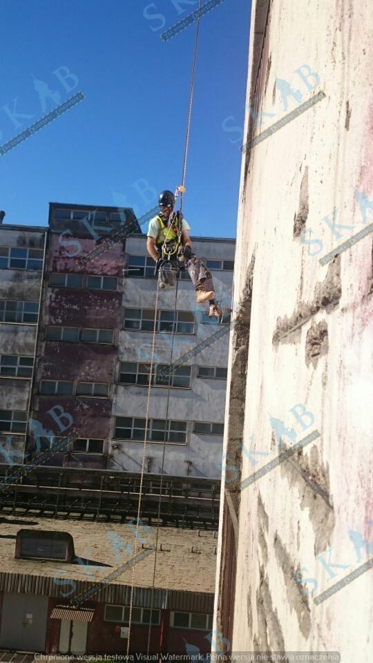 Pracownik na linie, zabezpieczony uprzężą, w trakcie pracy na wysokości przy ścianie budynku, widoczne uszkodzenia elewacji i budynek w tle.