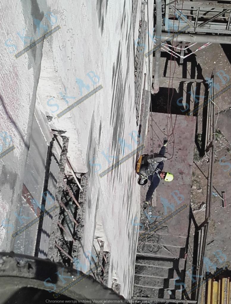 Pracownik w kasku, zabezpieczony linami, podczas renowacji elewacji budynku w Toruniu. Widok z góry na robotnika wiszącego na linie przy ścianie z widocznymi uszkodzeniami.