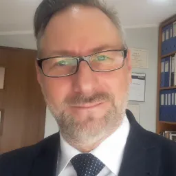 Portret uśmiechniętego mężczyzny w okularach, garniturze i krawacie w biurze, z dyplomami i segregatorami w tle.