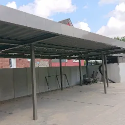 Nowoczesna, metalowa konstrukcja zadaszenia, prawdopodobnie carport, z widocznymi elementami montażowymi i narzędziami, na tle ceglanego budynku i pochmurnego nieba.