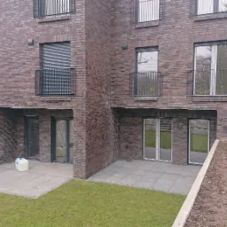 Nowoczesny budynek z ciemnej cegły z balkonami z prostymi, metalowymi balustradami, widok na trawnik i elementy wyposażenia budowlanego.