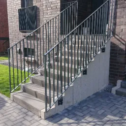 Zewnętrzne schody betonowe z czarną metalową balustradą przed budynkiem z cegły klinkierowej, widok z dołu na kostkę brukową.