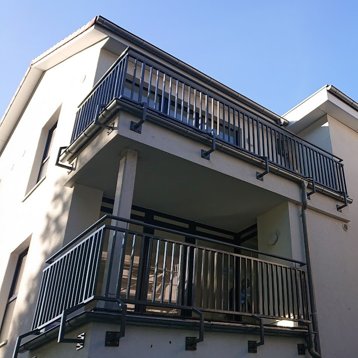 Dwupoziomowy balkon z szarymi, metalowymi balustradami o pionowych szczebelkach, zamontowanymi na elewacji budynku w słoneczny dzień.