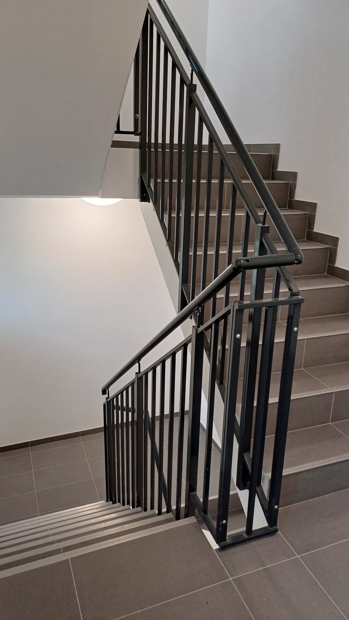 Klatka schodowa z szarymi płytkami i czarną, metalową balustradą z pionowymi szczeblami, widok z góry, oświetlenie sufitowe.