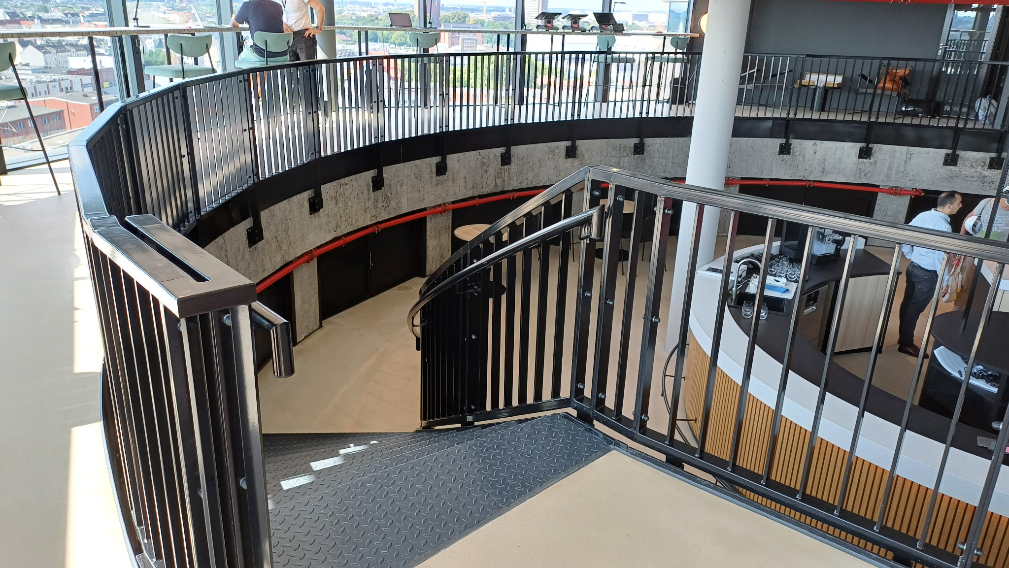 Wielopoziomowe wnętrze z czarnymi metalowymi balustradami, spiralnymi schodami z blachy ryflowanej i widokiem na miasto w tle.