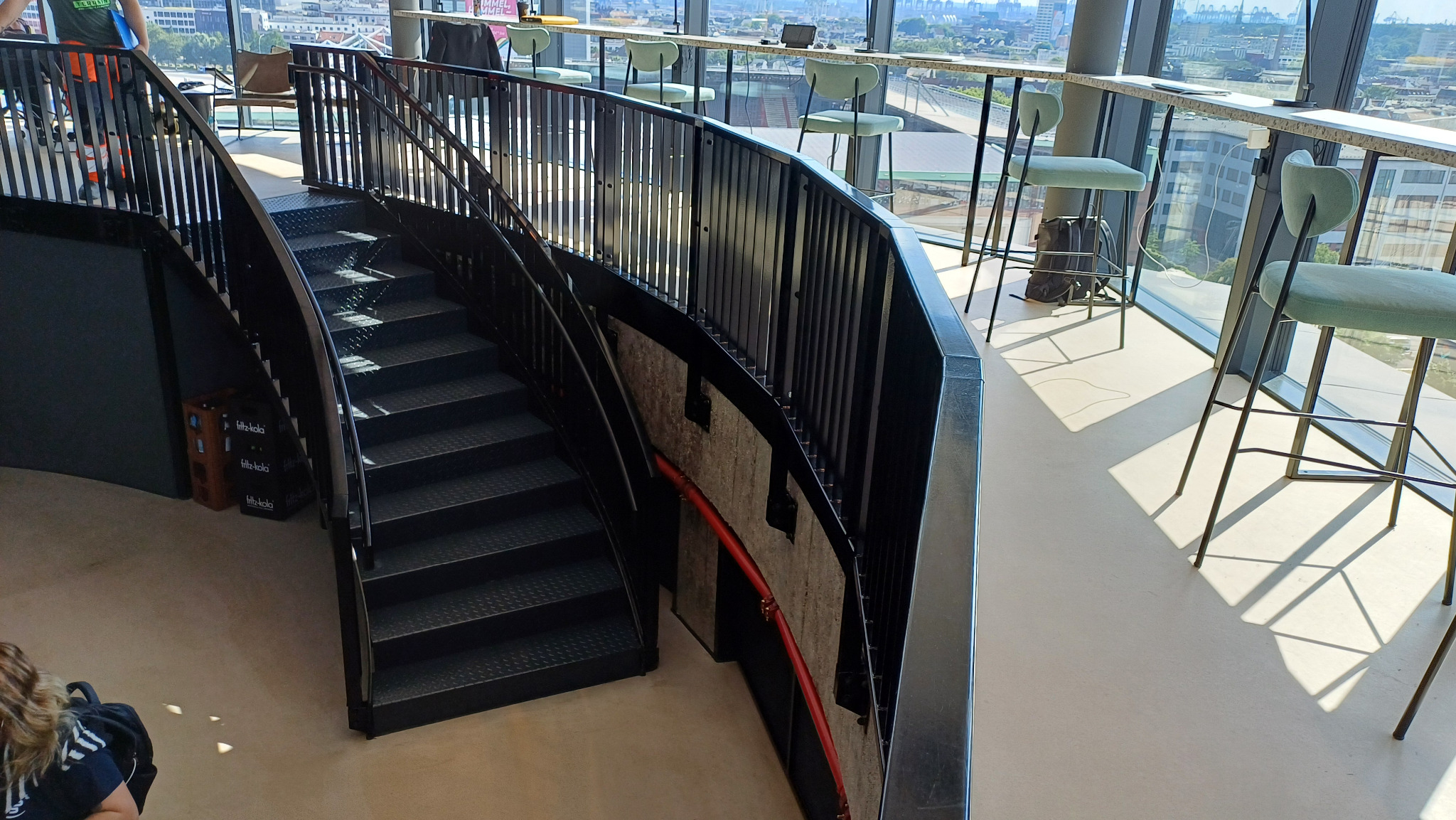 Wewnętrzne, metalowe schody spiralne z czarną balustradą w nowoczesnym budynku z widokiem na miasto przez panoramiczne okna, obok wysokie krzesła barowe przy blacie.