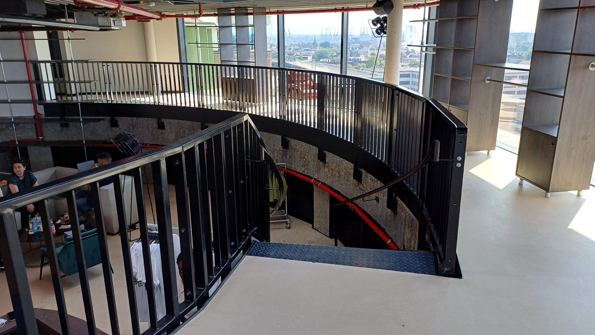 Wnętrze loftu z metalowymi schodami i balustradami, widok na miasto przez duże okna, betonowe ściany i czerwone rury instalacyjne.