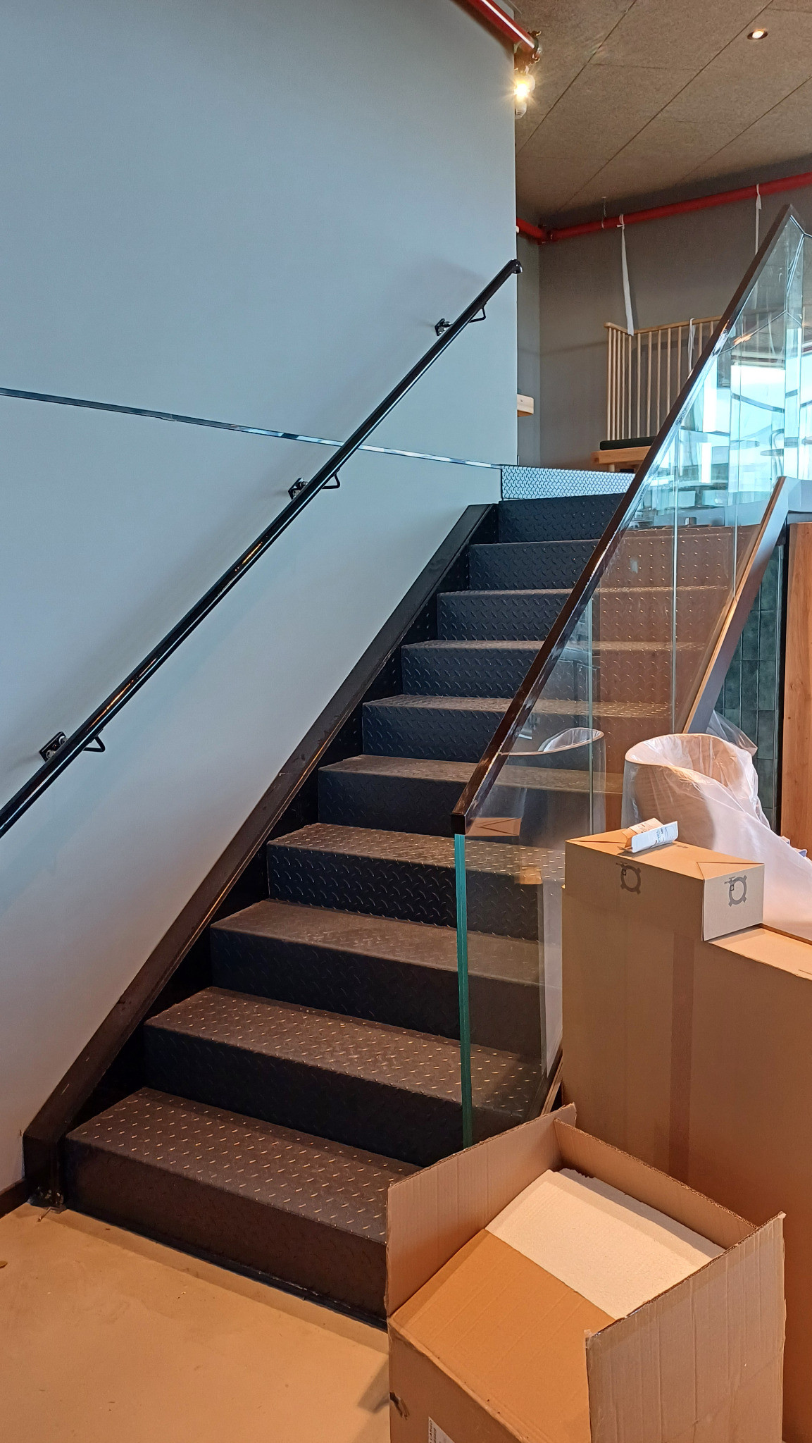 Metalowe schody z czarnymi stopniami o fakturze rombów i szklaną balustradą, widoczne kartony i folia ochronna, sugerujące świeży montaż.