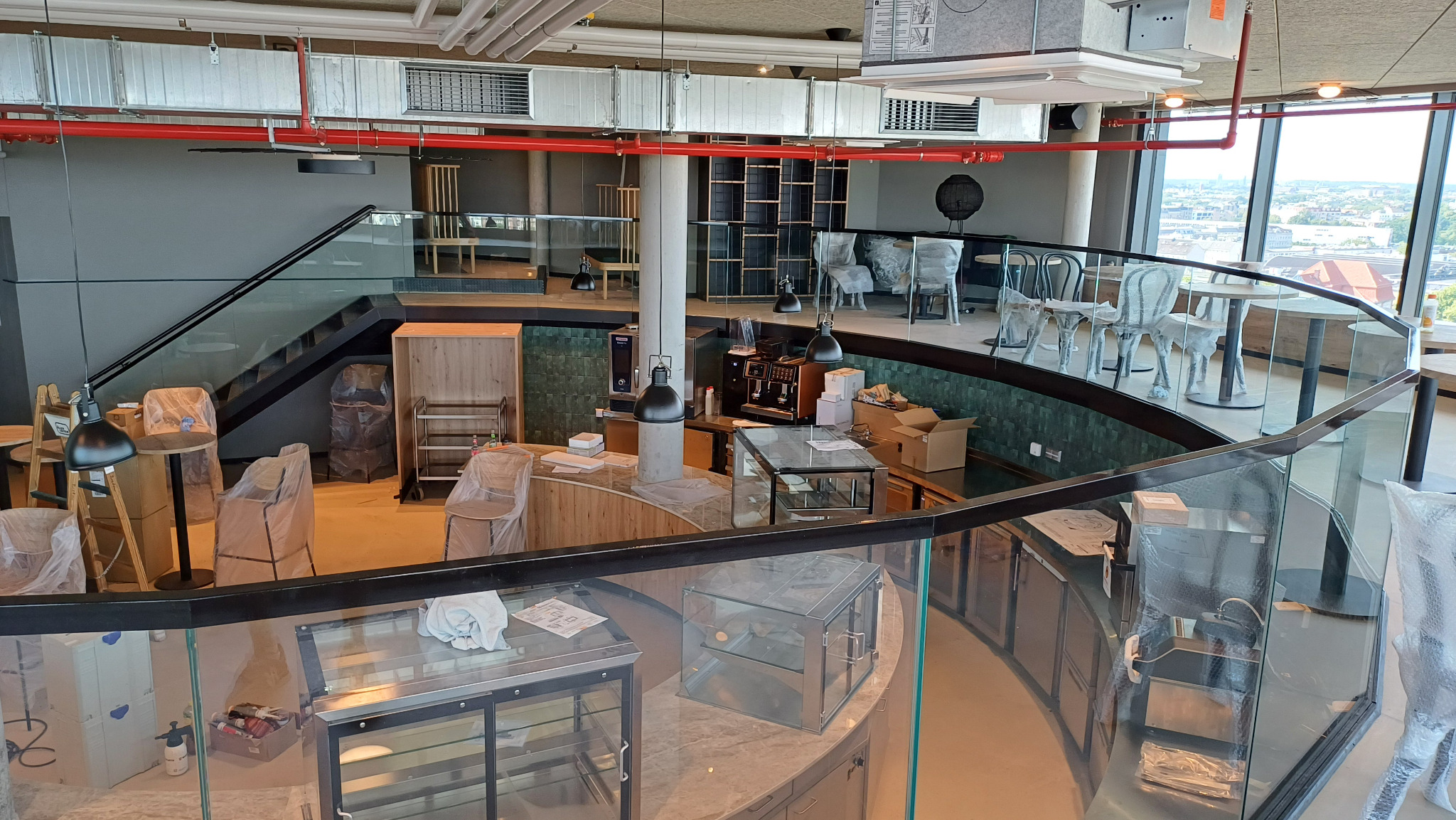 Wnętrze lokalu usługowego w trakcie wykańczania: widoczne szklane balustrady, schody, przygotowywany bar z wyposażeniem, meble zabezpieczone folią, duże okna z widokiem na miasto.