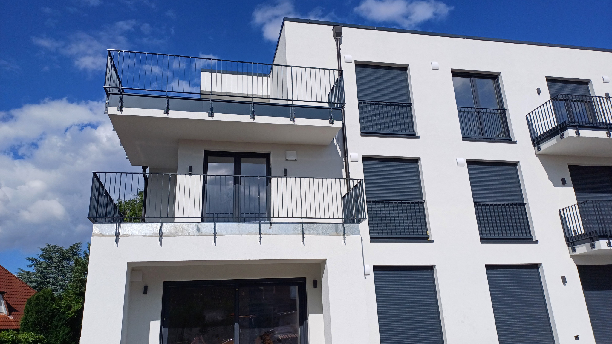 Nowoczesny budynek wielorodzinny z białymi elewacjami i grafitowymi balustradami balkonowymi oraz roletami zewnętrznymi, widok z dołu na błękitne niebo z chmurami.