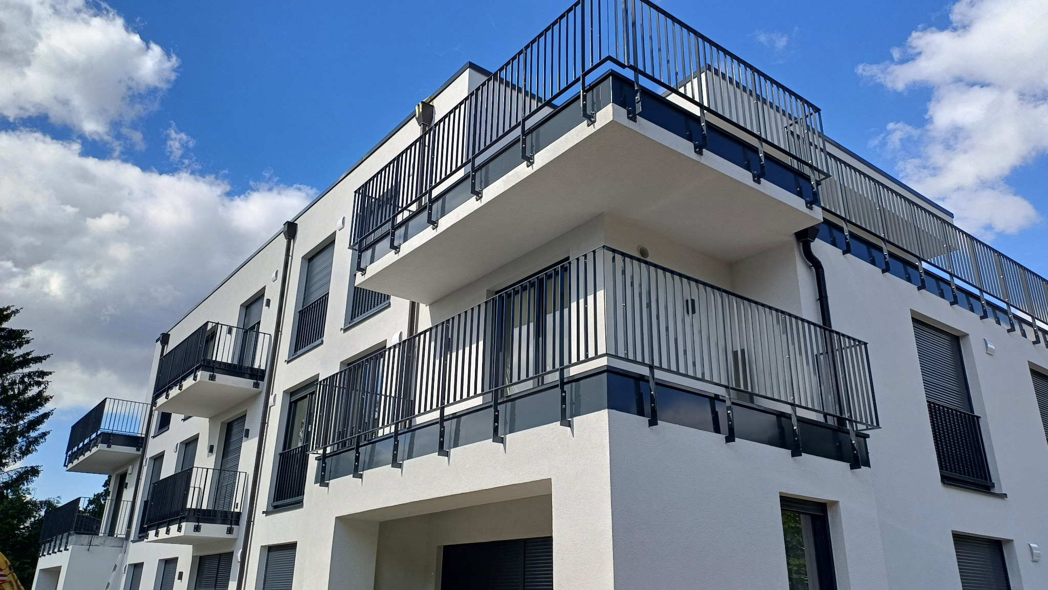 Nowoczesny budynek wielorodzinny z białymi ścianami i czarnymi balustradami balkonowymi z elementami szkła, widok z dołu na tle błękitnego nieba z chmurami.