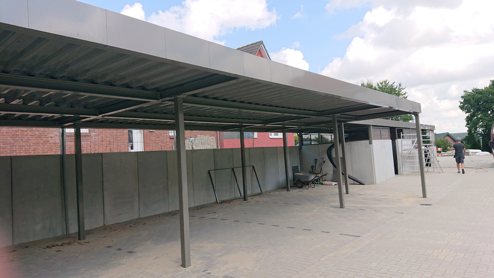 Nowoczesna, metalowa konstrukcja zadaszenia, prawdopodobnie carport, z widocznymi elementami montażowymi i narzędziami, na tle ceglanego budynku i pochmurnego nieba.