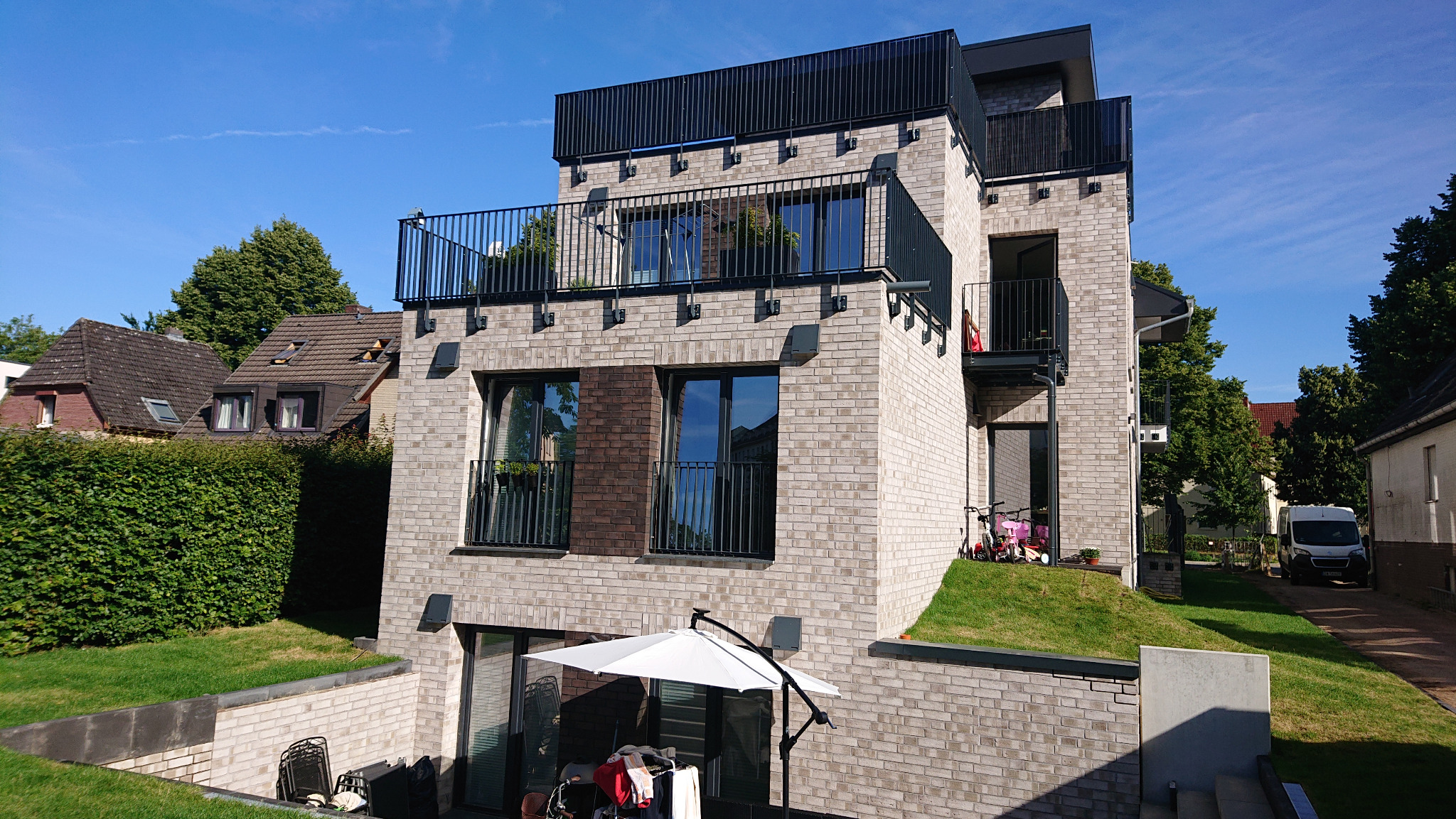 Nowoczesny dom z jasnej cegły z balkonami i czarnymi balustradami, widok z poziomu niższego parteru, z fragmentem ogrodu i parasolem.