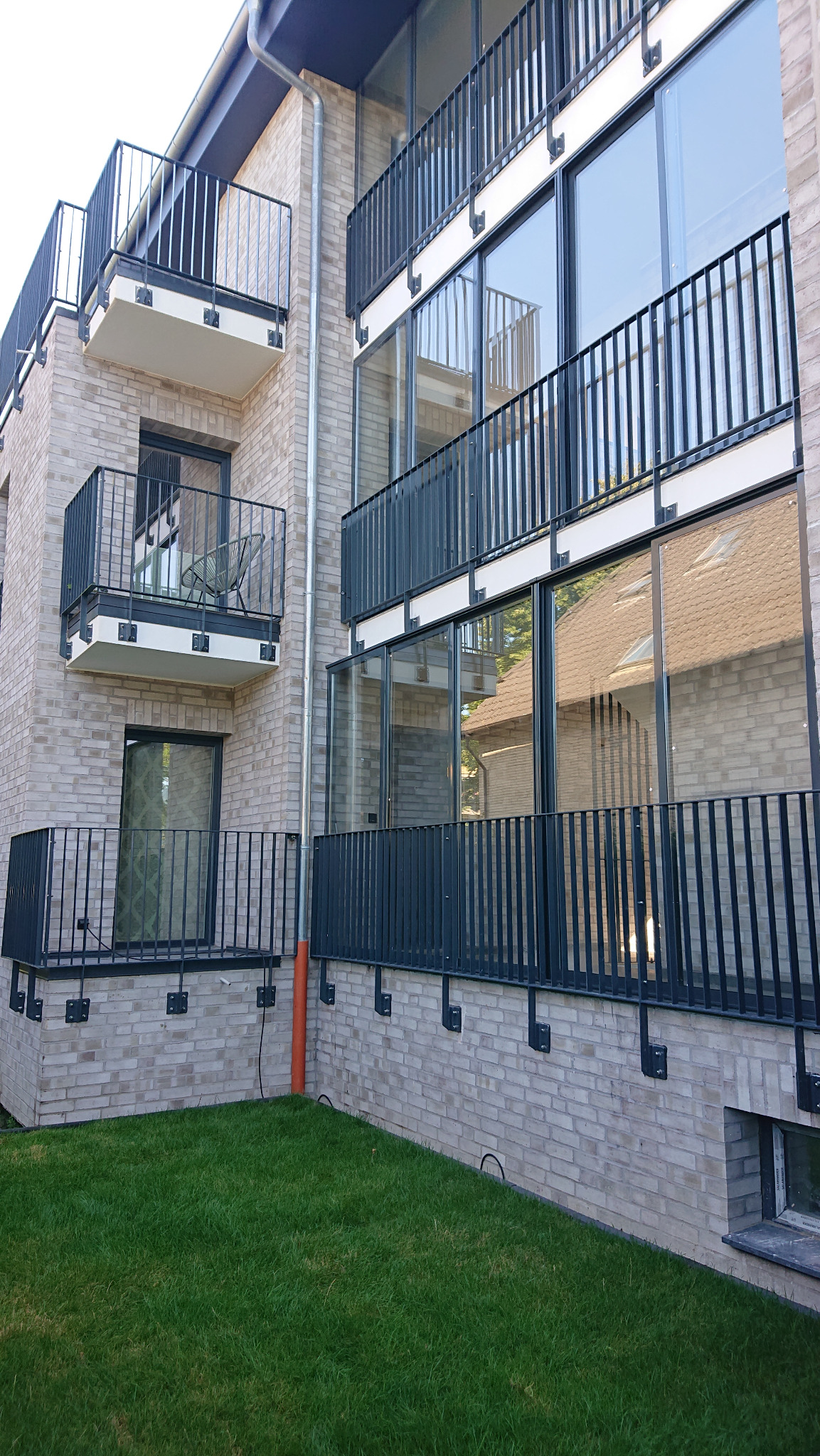 Nowoczesny budynek z jasnej cegły z balkonami z ciemnymi, metalowymi balustradami i dużymi oknami, widok z poziomu trawnika.