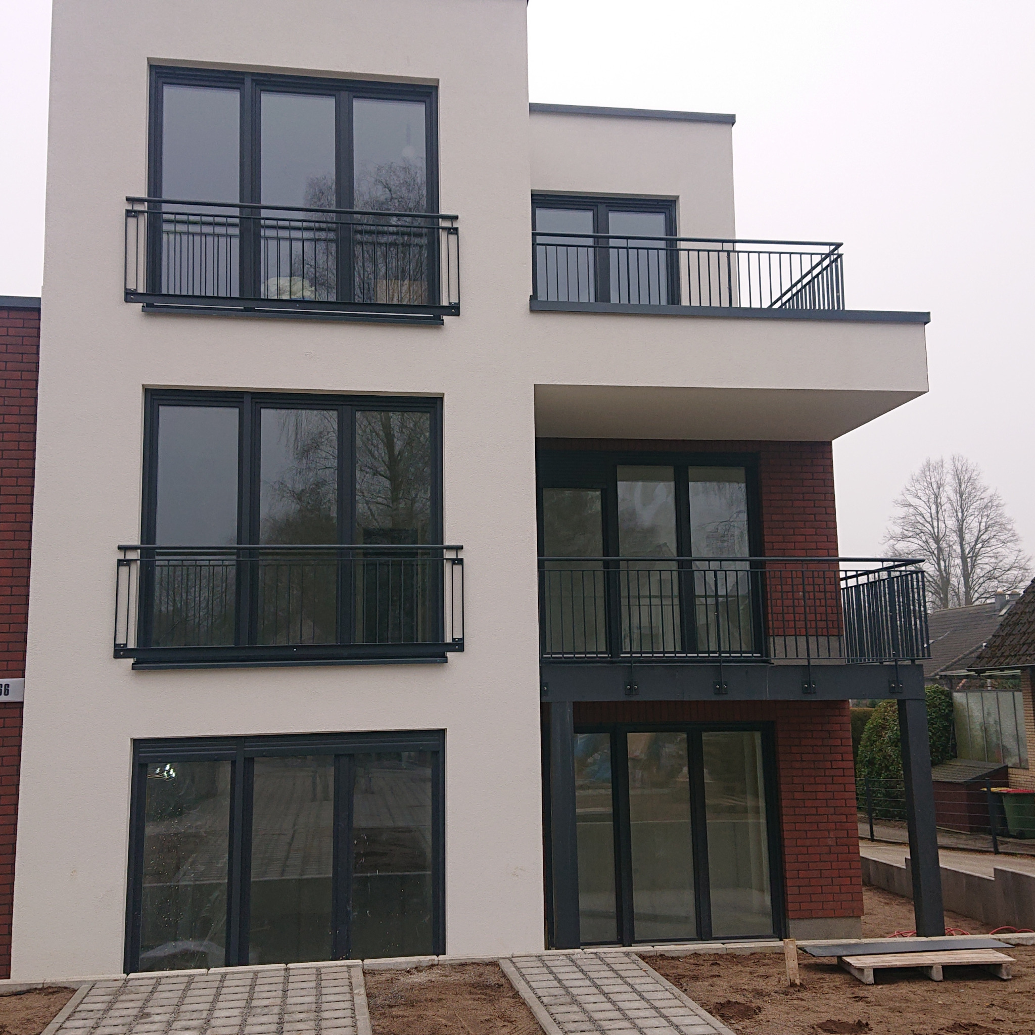 Nowoczesny budynek wielorodzinny z balkonami z czarnymi, metalowymi balustradami, elewacja w kolorze złamanej bieli z elementami z czerwonej cegły, widok z poziomu gruntu na trzy kondygnacje.