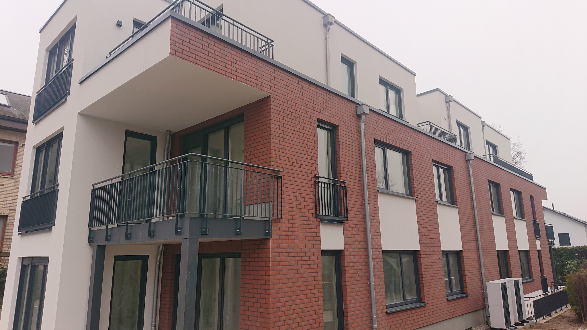Nowoczesny budynek wielorodzinny z czerwonej cegły z balkonami z czarnymi, metalowymi balustradami, fragment ocieplonej elewacji oraz widoczne jednostki klimatyzacyjne na zewnątrz.