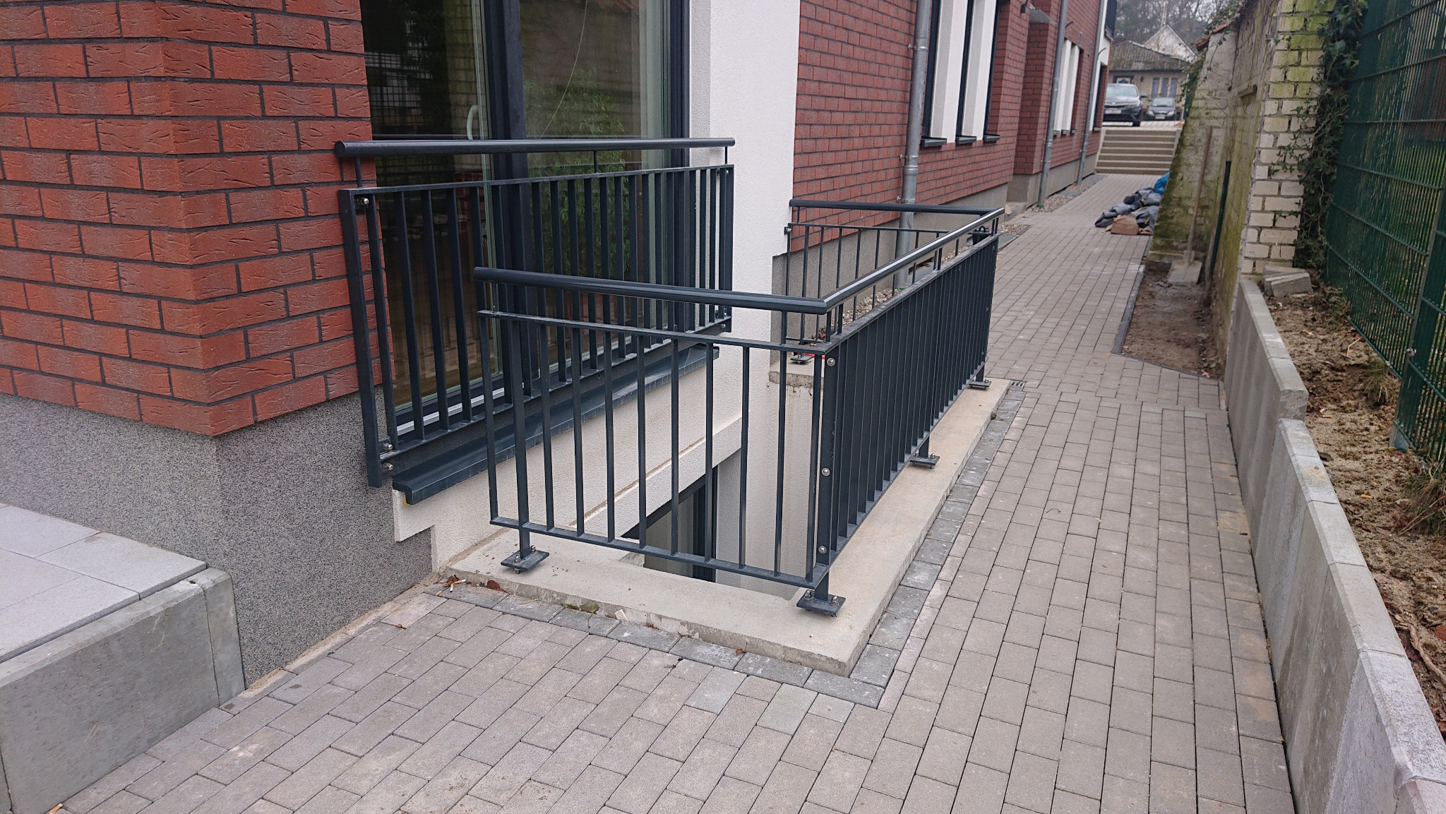 Metalowa balustrada zabezpieczająca zejście do piwnicy przy budynku z czerwonej cegły, widok z zewnątrz na chodnik z kostki brukowej i sąsiednią posesję.