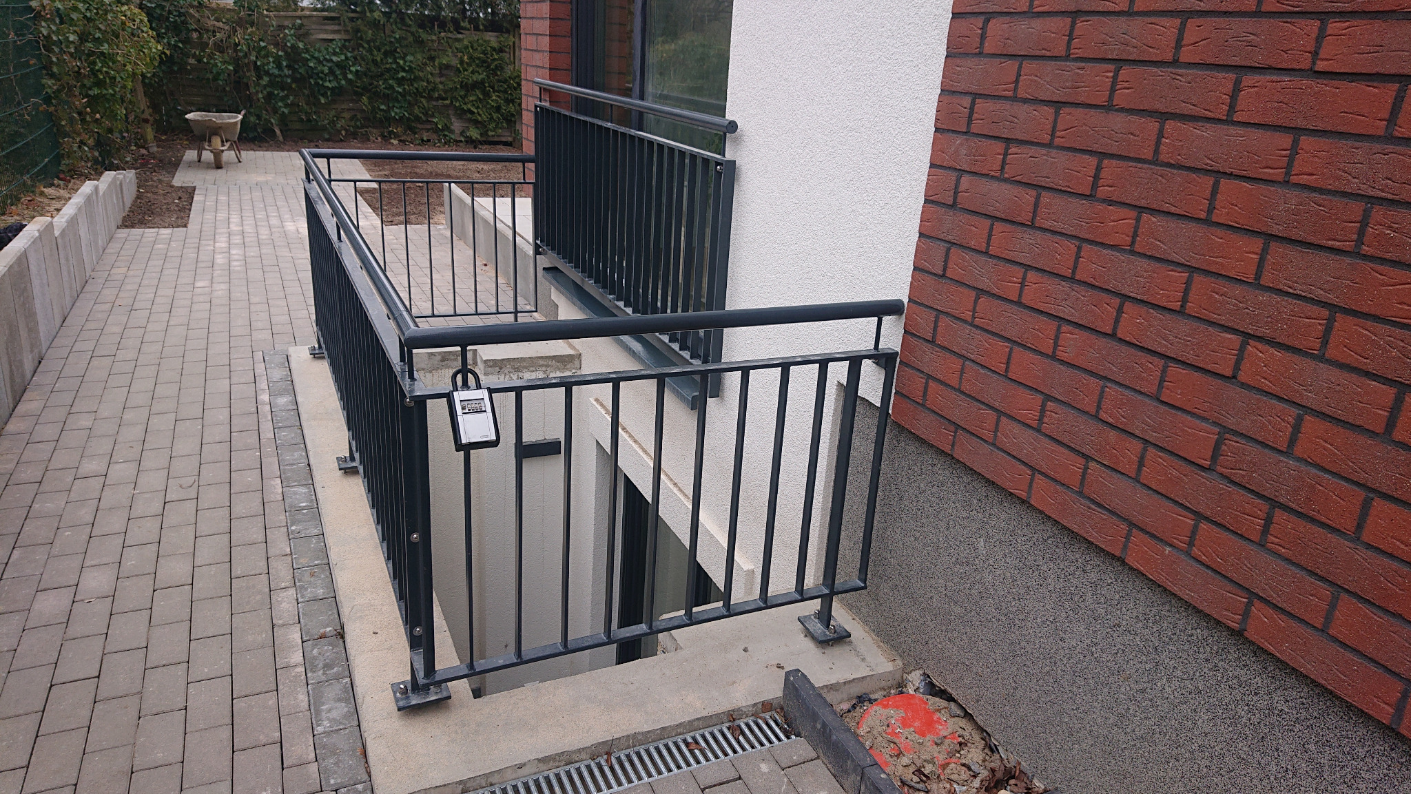 Metalowa balustrada zabezpieczająca zejście do piwnicy, z cyfrowym zamkiem na jednym z paneli, obok elewacja wykończona czerwoną cegłą klinkierową.