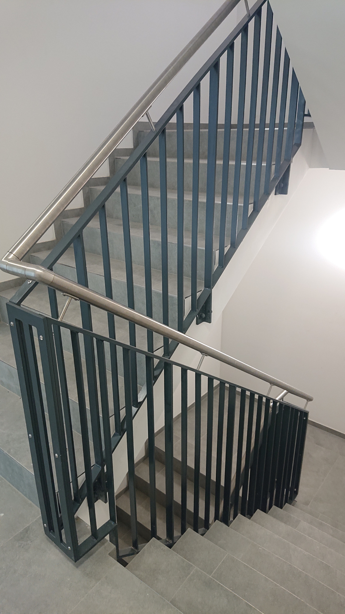 Szare schody z metalową, ciemną balustradą z pionowymi szczebelkami i nierdzewną poręczą, widok z góry.