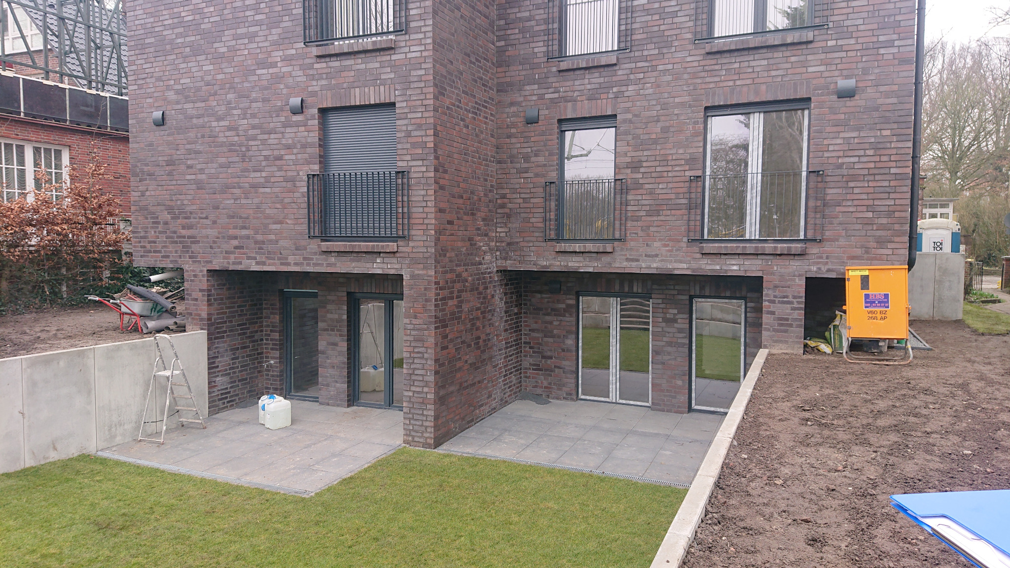 Nowoczesny budynek z ciemnej cegły z balkonami z prostymi, metalowymi balustradami, widok na trawnik i elementy wyposażenia budowlanego.