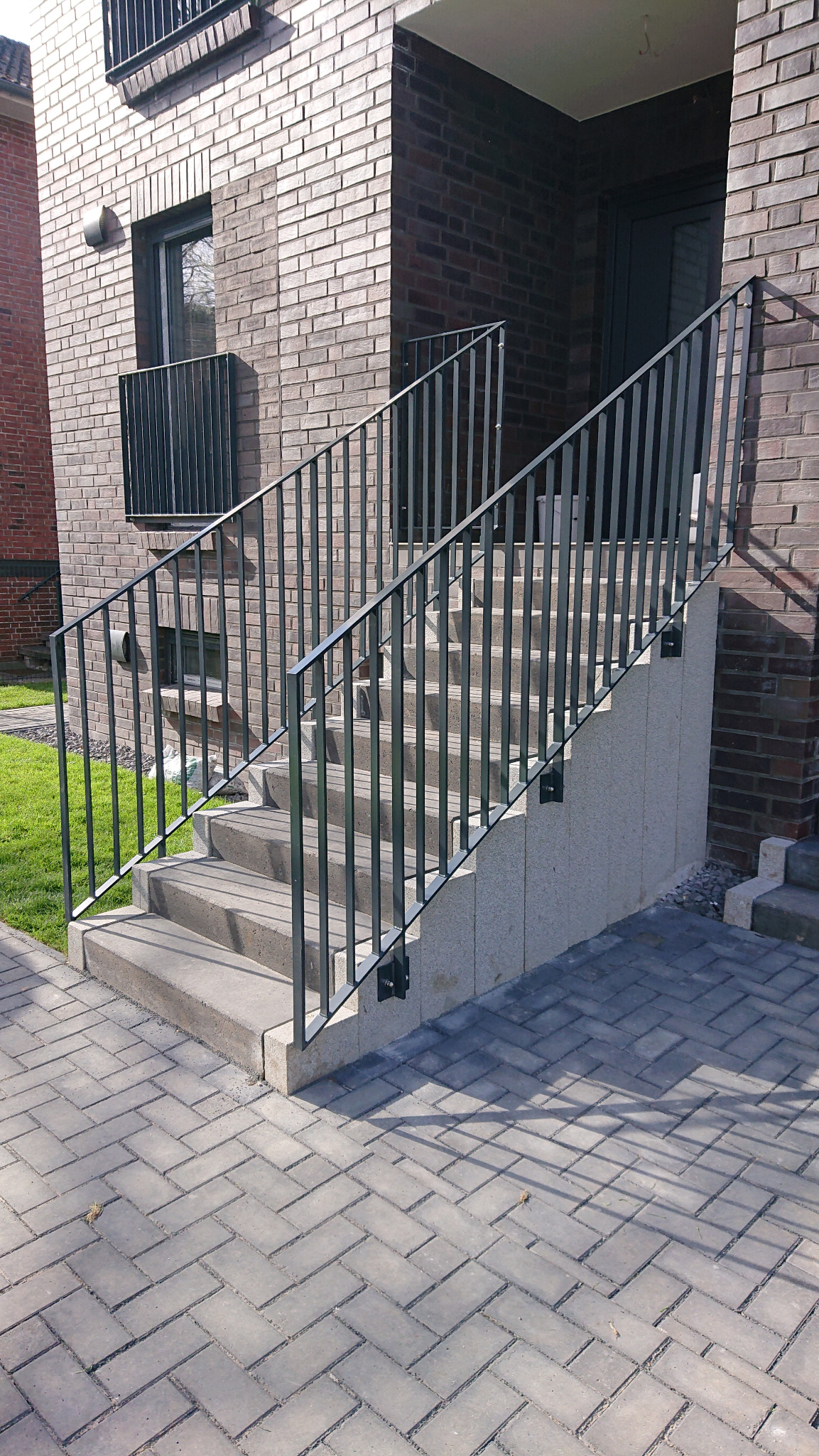 Zewnętrzne schody betonowe z czarną metalową balustradą przed budynkiem z cegły klinkierowej, widok z dołu na kostkę brukową.