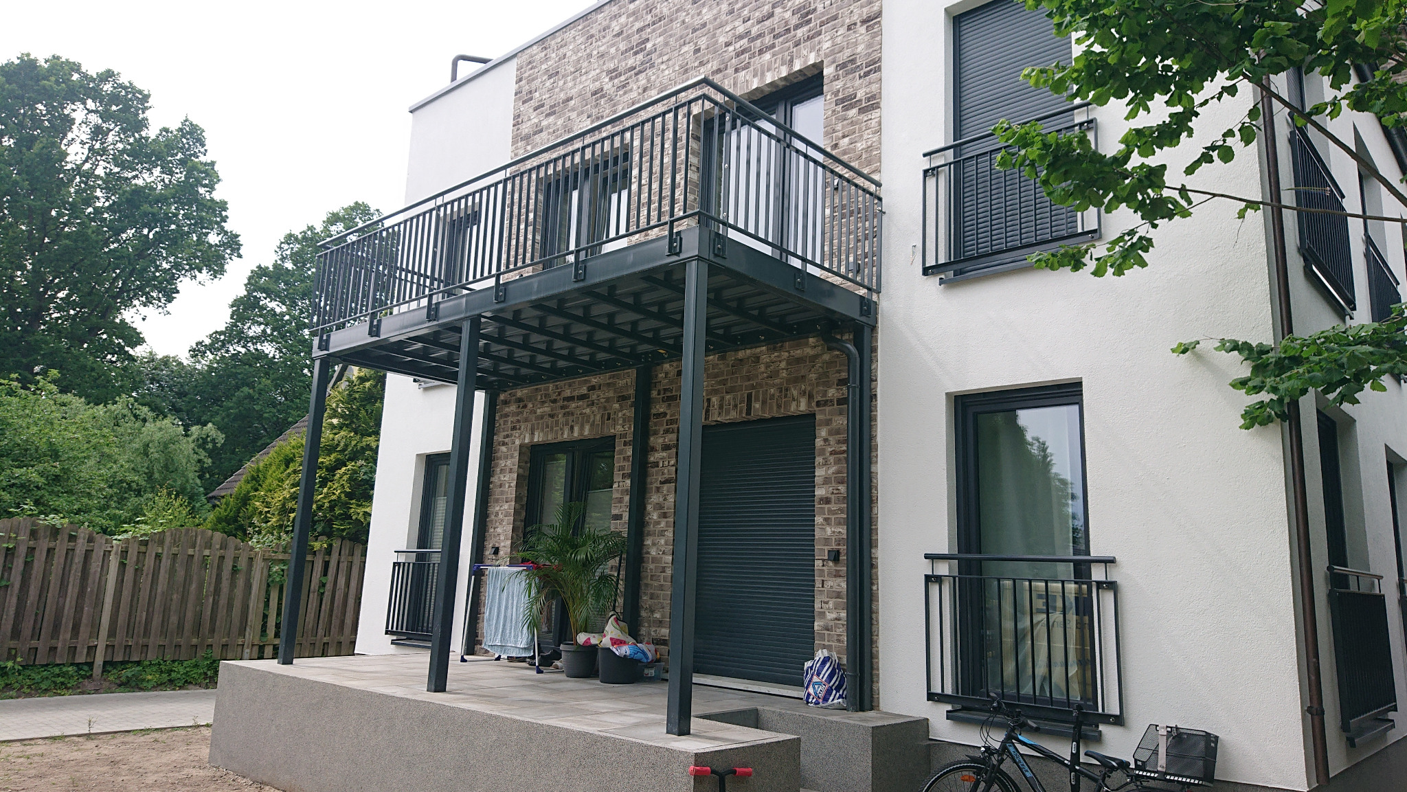 Nowoczesny budynek z balkonami z czarnymi, metalowymi balustradami, z widocznymi roślinami na parterze i rowerem zaparkowanym obok.