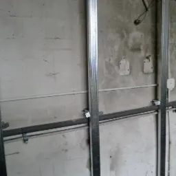Surowa ściana z widocznymi metalowymi profilami konstrukcyjnymi, rurami instalacyjnymi i puszką elektryczną, przygotowana do dalszych prac wykończeniowych.
