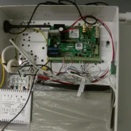 Otwarta skrzynka z elektroniką alarmową: centrala alarmowa z kartą SIM, transformator AWT050 20VA, akumulator w folii i okablowanie w białej obudowie.