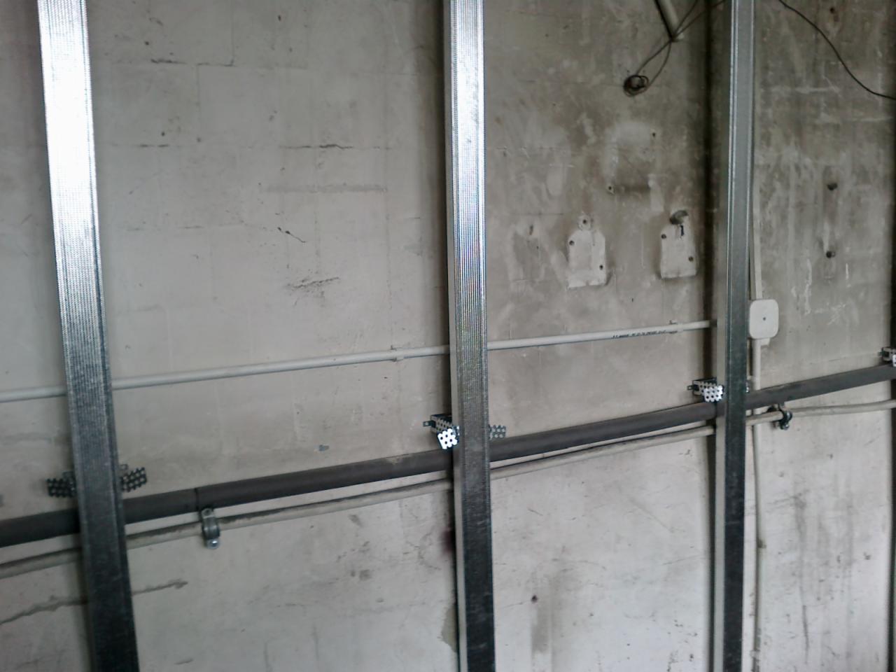 Surowa ściana z widocznymi metalowymi profilami konstrukcyjnymi, rurami instalacyjnymi i puszką elektryczną, przygotowana do dalszych prac wykończeniowych.