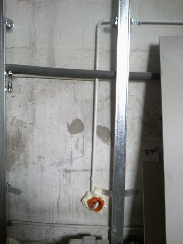 Instalacja elektryczna w trakcie montażu: przewód w rurce PCV wychodzący ze ściany, zabezpieczony pianką montażową wokół pomarańczowej puszki, widoczne profile metalowe konstrukcji ściany działowej.