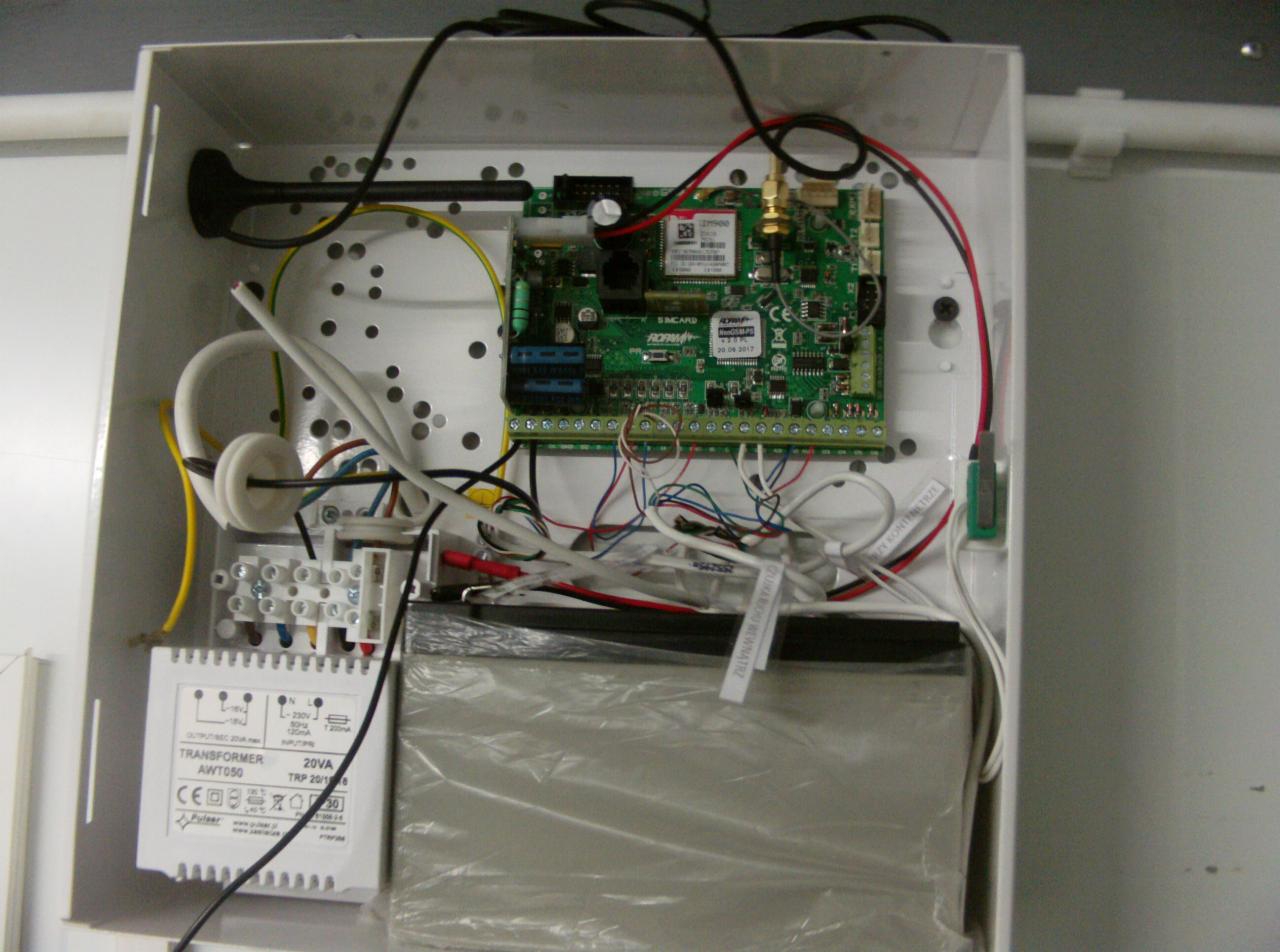 Otwarta skrzynka z elektroniką alarmową: centrala alarmowa z kartą SIM, transformator AWT050 20VA, akumulator w folii i okablowanie w białej obudowie.
