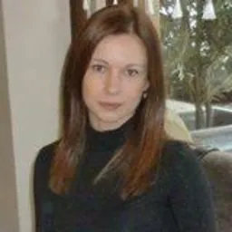 Portret kobiety o brązowych włosach, w czarnym golfie, na rozmytym tle wnętrza z oknem. Ujęcie od przodu, neutralny wyraz twarzy.