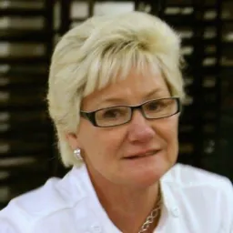 Portret kobiety w okularach, z blond włosami i srebrną biżuterią, ubrana w białą koszulę, na rozmytym tle.