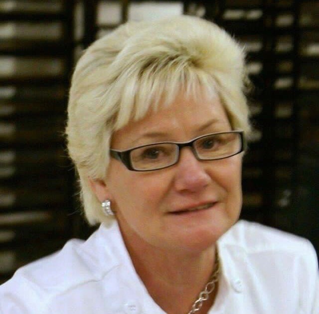 Portret kobiety w okularach, z blond włosami i srebrną biżuterią, ubrana w białą koszulę, na rozmytym tle.