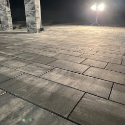 BRUK-OGRODY BARTOSZ MIKOŁAJEWSKI - Ułożona szara kostka brukowa w nocnym oświetleniu reflektorami, widoczne słupki z numerem domu.