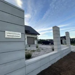 Nowoczesne ogrodzenie modułowe z szarego betonu architektonicznego z wbudowaną skrzynką na listy, na tle domu jednorodzinnego i błękitnego nieba z chmurami.