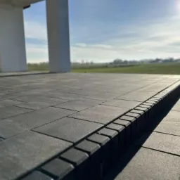 Wykończony taras z ciemnoszarej kostki brukowej o regularnym kształcie, z obrzeżem z kostki ustawionej pionowo, na tle białego domu i zielonego krajobrazu w słoneczny dzień.