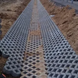 Układanie ażurowych płyt betonowych na przygotowanym podłożu, widoczny pracownik w kasku i kamizelce odblaskowej, teren budowy z ziemią po bokach.