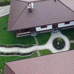 Widok z góry na dom z brązowym dachem, okrągły podjazd i ścieżki z jasnej kostki, zadbany trawnik i rabaty kwiatowe.