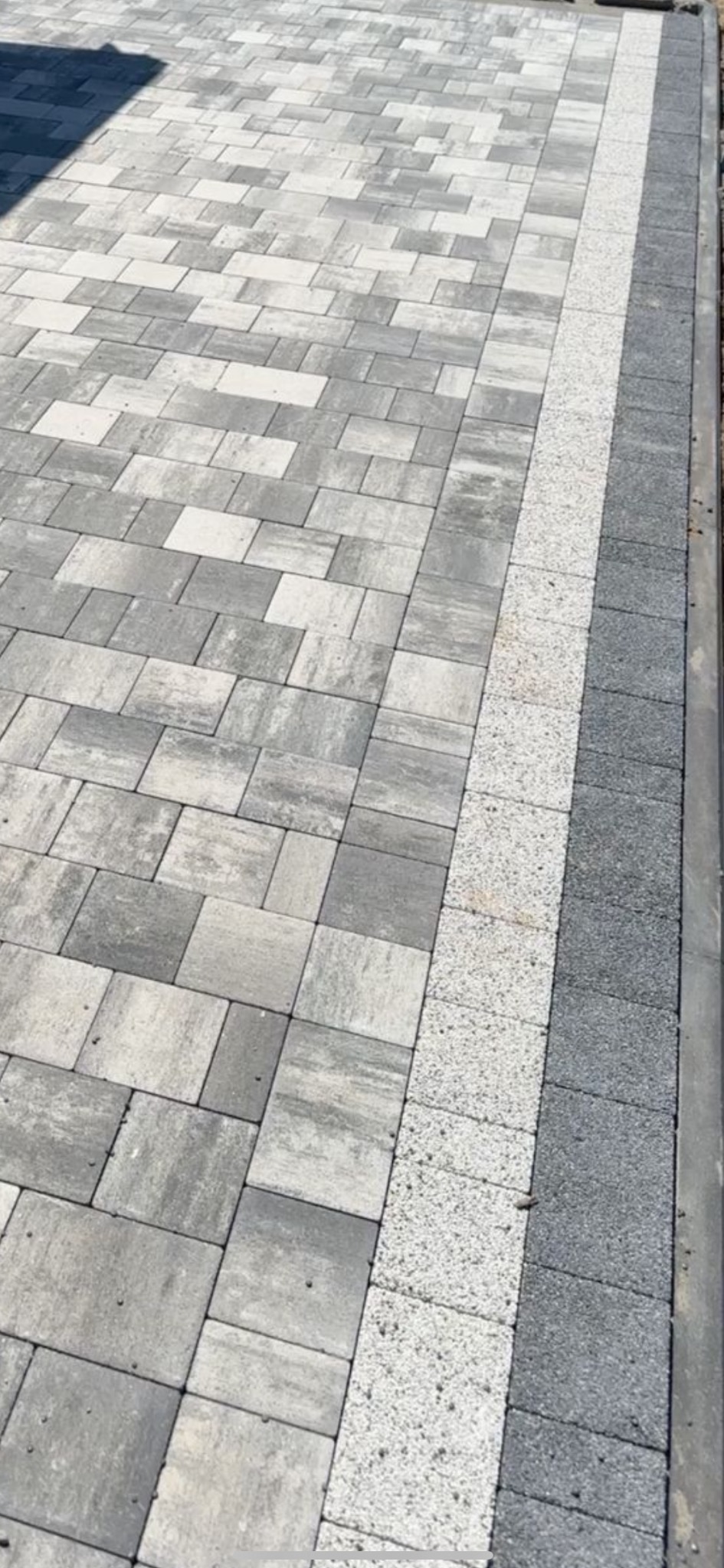 Świeżo ułożona nawierzchnia z kostki brukowej w odcieniach szarości, z dekoracyjnym pasem z jasnego kruszywa i ciemnoszarym obrzeżem.