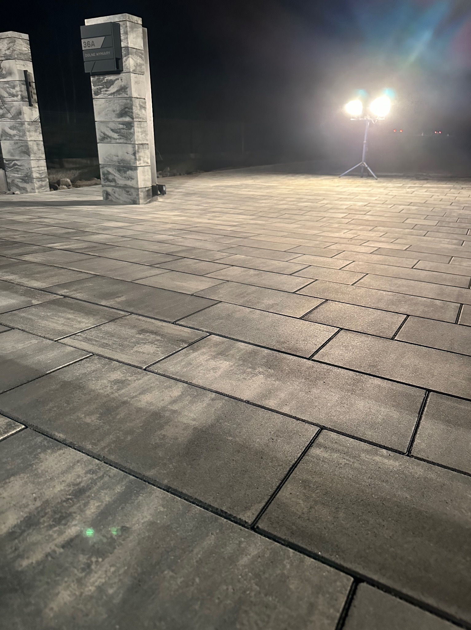 Ułożona szara kostka brukowa w nocnym oświetleniu reflektorami, widoczne słupki z numerem domu.