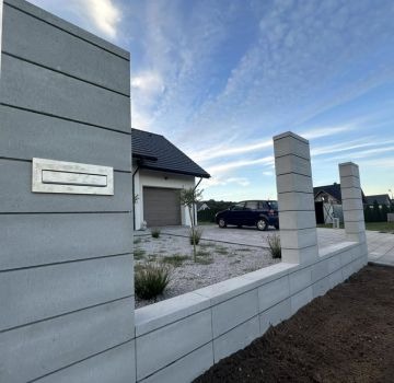 Nowoczesne ogrodzenie modułowe z szarego betonu architektonicznego z wbudowaną skrzynką na listy, na tle domu jednorodzinnego i błękitnego nieba z chmurami.