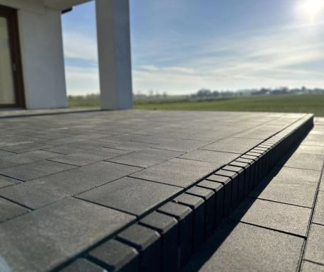 Wykończony taras z ciemnoszarej kostki brukowej o regularnym kształcie, z obrzeżem z kostki ustawionej pionowo, na tle białego domu i zielonego krajobrazu w słoneczny dzień.