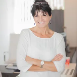 Uśmiechnięta brunetka w białej bluzce, z założonymi rękami, w jasnym biurze. Na głowie okulary, widoczny zegarek na ręku. Portret biznesowy.