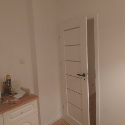 MARAND MARCIN JACH - Świeżo pomalowane na biało ściany i drzwi wewnętrzne z poziomymi frezami, widoczne narzędzia malarskie na szafce i drewniana podłoga.