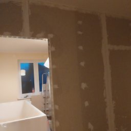 MARAND MARCIN JACH - Ściana w trakcie szpachlowania, widoczne łączenia płyt gipsowo-kartonowych i punktowe uzupełnienia ubytków, w tle okno i drabina.