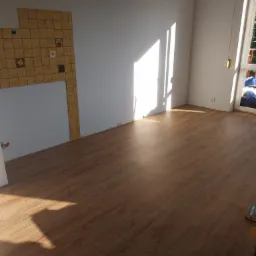 Pusty pokój po remoncie z nową, brązową podłogą, fragmentem ściany z niedokończonymi kafelkami i promieniami słońca wpadającymi przez okno.